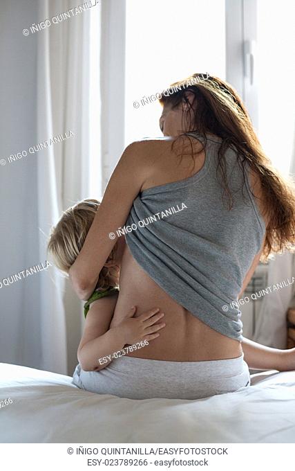 blonde caucasian baby two years old in brunette woman mother arms breastfeeding sitting on bed white clothes in bedroom next to window indoor