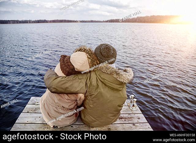 Back view of young couple in love sitting on a jetty looking at lake in winter