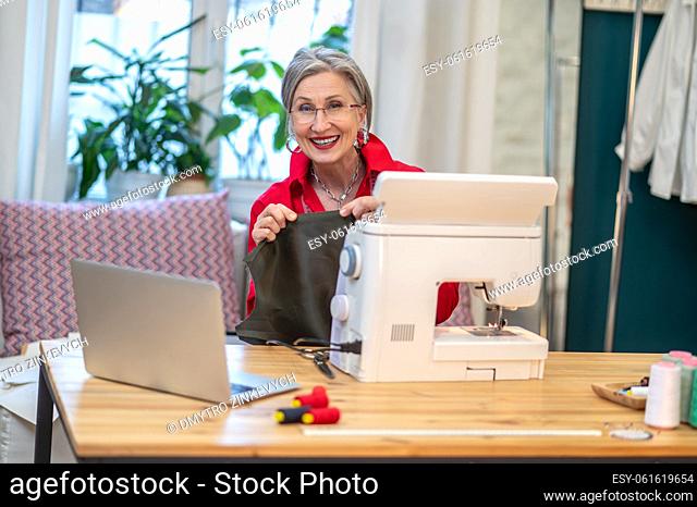 Lucky day. Friendly stylish woman in glasses holding fabric sitting at table with sewing machine and laptop smiling at camera