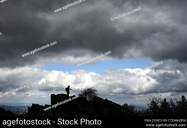 12 April 2021, Hessen, Schmitten: A hiker stands on the Brunhildis rock on the Großer Feldberg in the Taunus, over which dark clouds have gathered