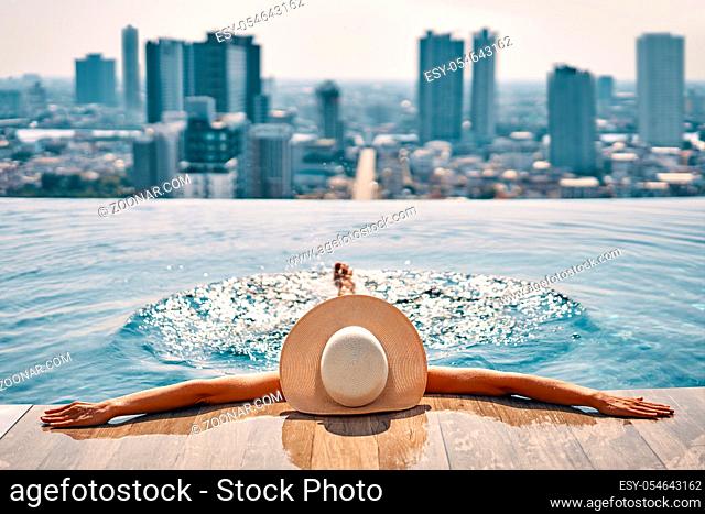 Back view of young woman in hat relaxing in swimming pool on the roof top of hotel and enjoy cityscape. Summer vacation, rest concept