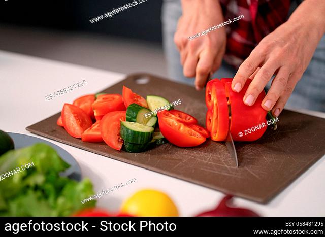 Cuts vegetables for a fresh salad young housewife cut bell pepper, cucumber, tomato on cutting board preparing for a family dinner standing in the new kitchen...