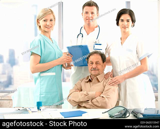 Portrait of senior patient sitting at desk, medical team around, smiling at camera