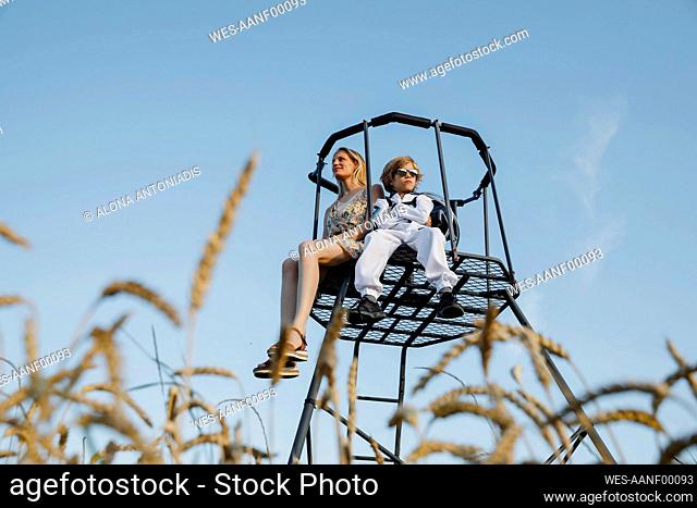 Son wearing space suit sitting with mother on lookout tower at field
