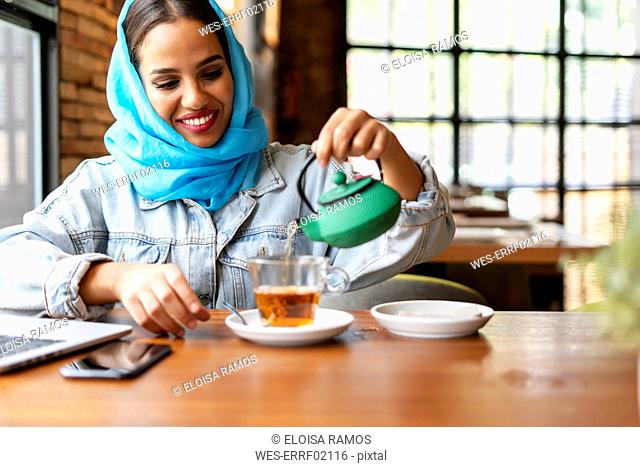 Businesswoman wearing turquoise hijab in a cafe and working, pouring tea