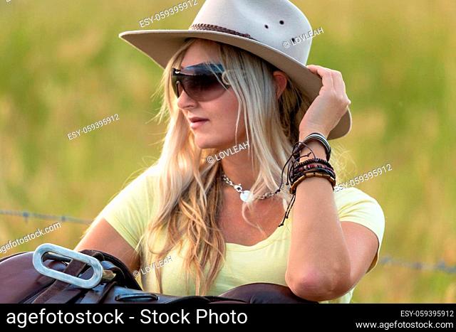 Casual dressed woman with akubra hat, oversized sunglasses leaning by her saddle. She is wearing leather and braided bangles