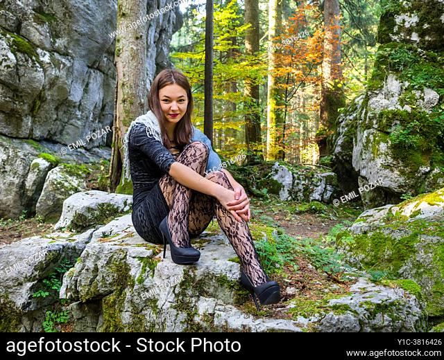 Young woman in nature sitting on big rock embracing knees patterned Black tights and stiletto pumps