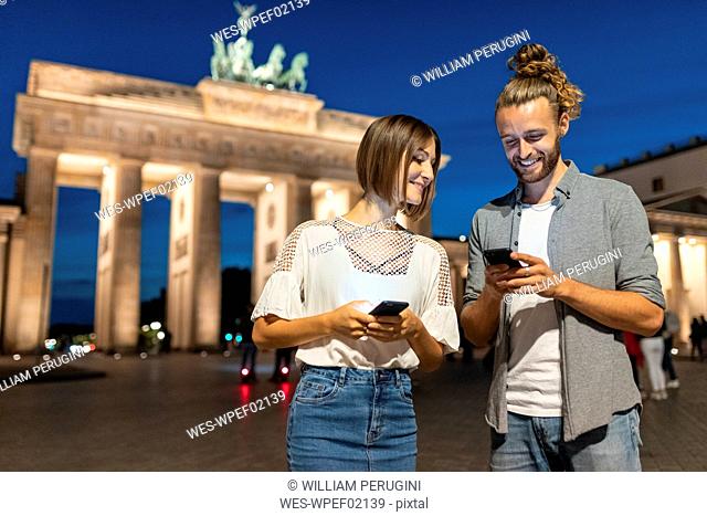 Happy couple using smartphones at Brandenburg gate at night, Berlin, Germany
