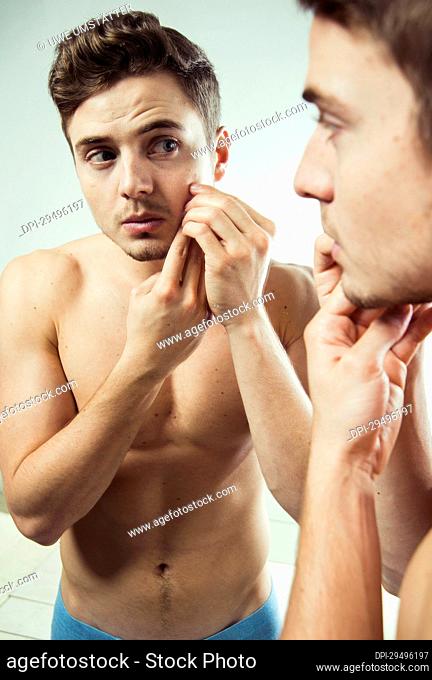 Close-up of young man looking at reflection in bathroom mirror and examining skin on face, studio shot on white background