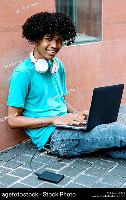 Smiling young man using laptop while sitting on footpath in city