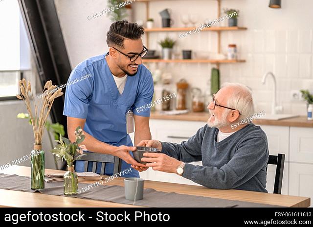 Cheerful amiable young caregiver giving a ceramic bowl of food to an elderly man seated at the kitchen table