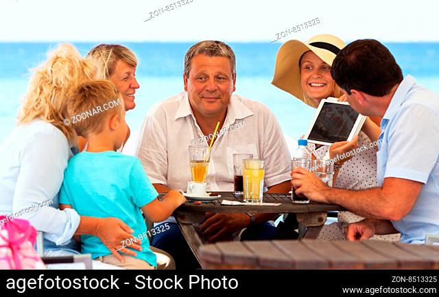 Woman with tablet computer showing photos to her big happy family. They spending summer day in outdoor cafe on the beach, sea view in background