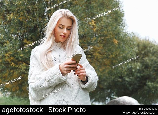 Portrait of young caucasian blonde hair woman in white pullover uses the phone outdoor