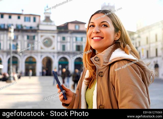 Copyspace photo of smiling young woman holding telephone with her hand wearing winter coat standing pensively looking up over city blurred background