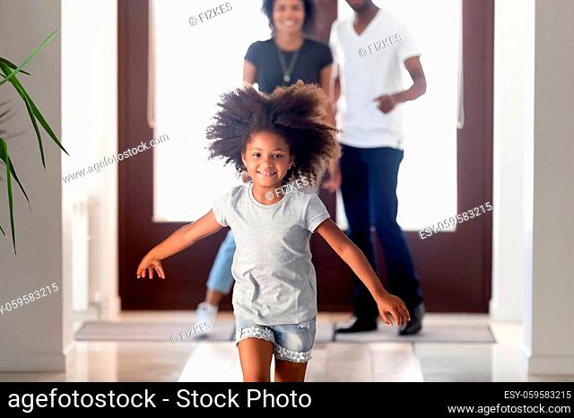 Happy african family and little girl arrived at new house, kid exploring new home feels excited running at hallway having fun indoors, parents looking at her