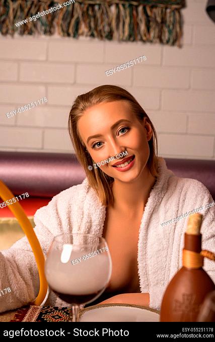 Young woman in bathrobe with hookah and wine glass on the table in sauna rest room interior view