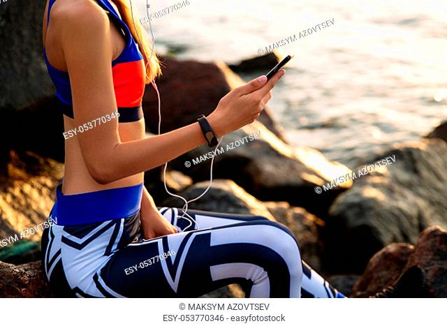 Unrecognized young woman in sportswear, in headset, using a mobile phone, sitting on stones, near the ocean