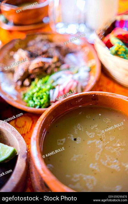 Close-up of clay bowl of lamb broth and plate of chopped lamb meat above orange and red tablecloth. Traditional soupy dish and condiments above colorful table