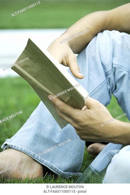 Young man sitting in grass, reading, close-up of low section
