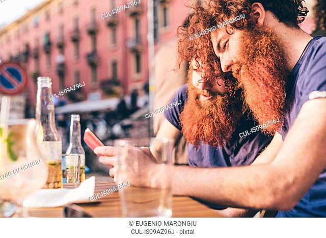 Young male hipster twins with red hair and beards reading smartphone texts at sidewalk bar