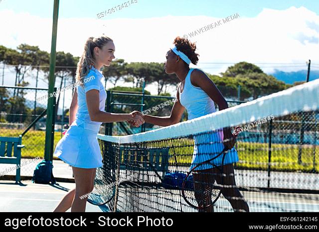 Confident young female multiracial competitors doing handshake over net at tennis court on sunny day