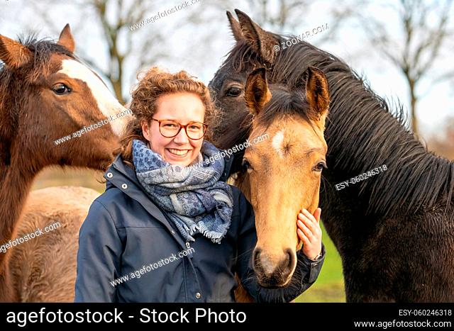 Smiling young woman in a wax coat with her 1 year old stallions in the pasture. She cuddles with them to socialize them. Three horse heads