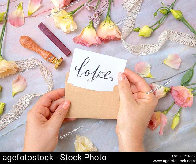 Hands with Card LOVE inside envelope near by pink flowers, wax seal and ribbons on a marble table top view. Romantic scene with handwritten card flat lay