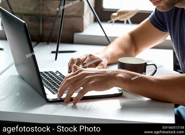 Bearded man is using a laptop on the blurry background of the window. Laptop is on the white wooden table with a cup near it