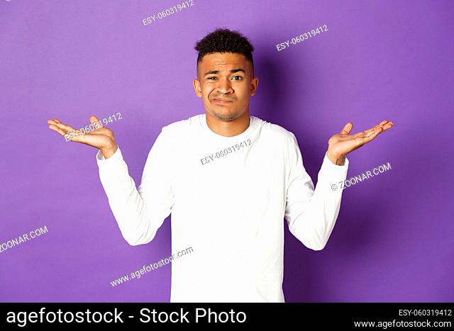 Portrait of indecisive african-american guy in white sweatshirt, raise hands sideways and shrugging clueless, standing confused against purple background