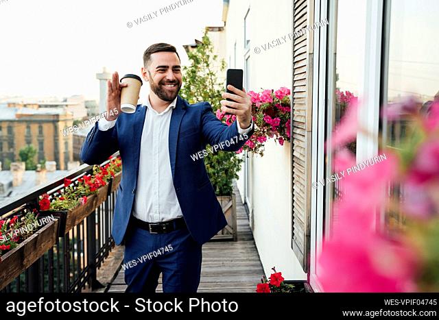 Smiling businessman waving during video call on smart phone in balcony