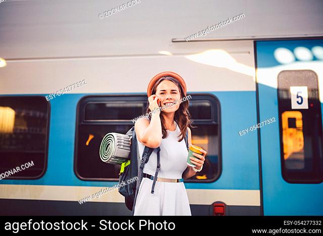 theme tourism and travel young student. beautiful young Caucasian girl in dress and hat standing at train station near train with backpack and cup coffee in...