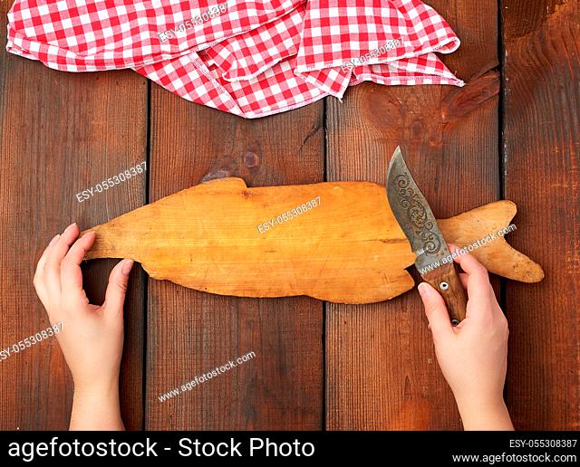 old empty wooden rectangular cutting board and hands hold a sharp knife, top view, brown wooden background