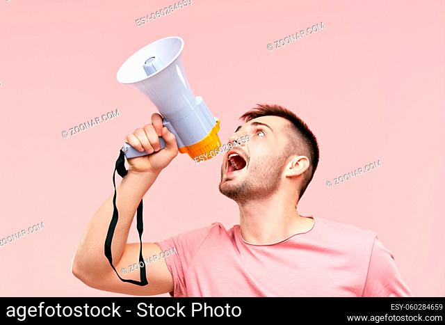 Funny young man shouting loud holding a megaphone over pink background. Announcement, communication concept