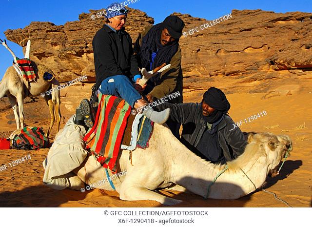 Tuareg nomads helping a female tourist to climb on a dromedary in the Acacous Mountains, Sahara desert, Libya
