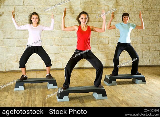 A group of young women exercising in the fitness club. They're smiling and looking at camera. They have hands up. Front view