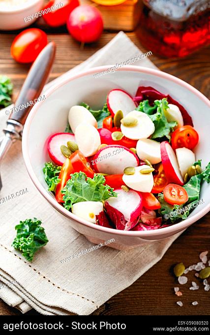 Salad from Iceberg lettuce, Kale and Radiccio, cherry tomatoes, radish, mini mozzarella and pumpkin seeds in bowls on dark wooden background