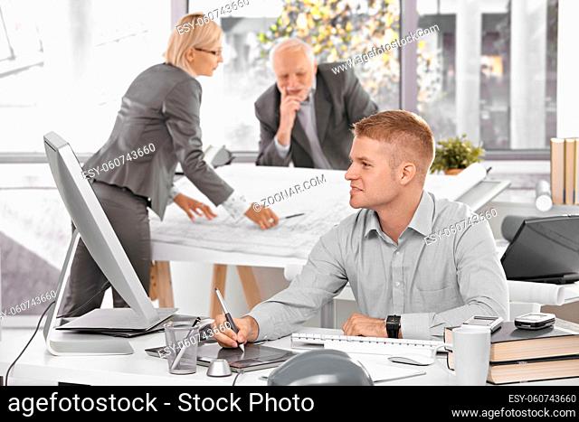 Designer team at work in office, young architect sitting at desk with drawing pad, older colleagues working on architectural plan