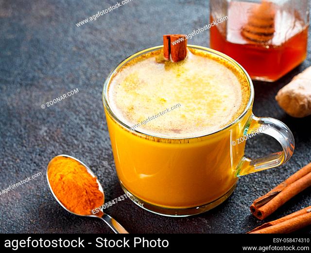 Healthy drink golden turmeric latte in glass cup.Gold milk with turmeric, ginger root, cinnamon sticks, turmeric powder and honey over black cement background