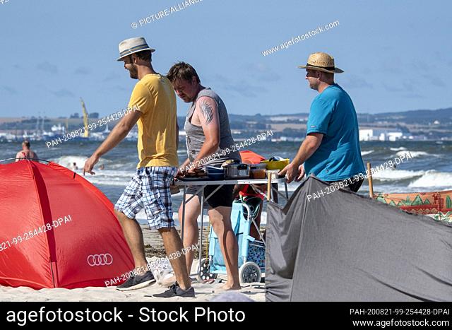 11 August 2020, Mecklenburg-Western Pomerania, Binz: Three men carry a table with pots, plates, bowls and food along the beach of the Baltic Sea