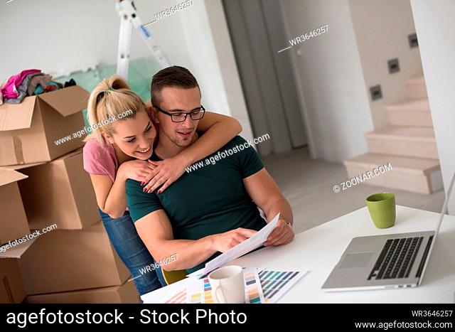 Young couple moving in a new home. Man and woman at the table using notebook laptop computer and plans with boxes around them