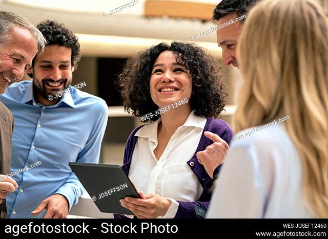 Happy businesswoman holding tablet PC discussing with colleagues in corridor