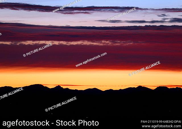 19 October 2021, Bavaria, Bernbeuren: The sky above the panorama of the Alps turns different shades of red shortly before sunrise