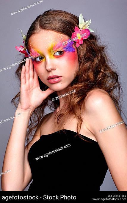 Portrait of teen girl with hand near face and orchid flower in wavy hair. Young female with unusual stylish make-up and false fashion feather eyelashes
