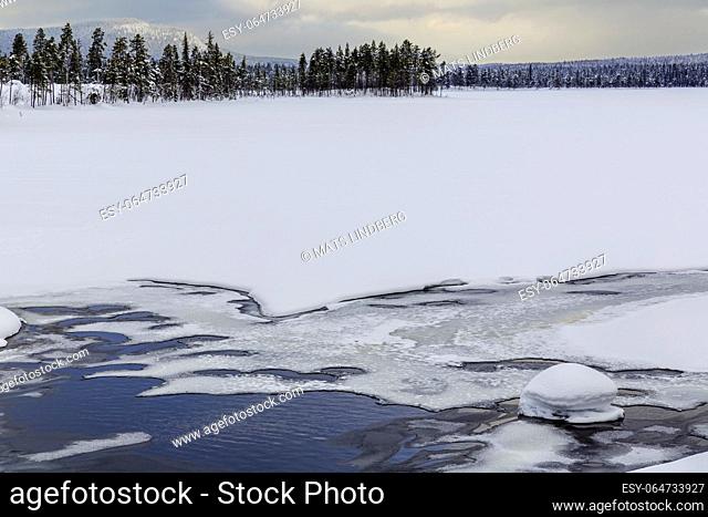 Winter landscape with frozen creek with partially open water, snowy trees and a mountain in background, Jokkmokk county, Swedish Lapland, Sweden