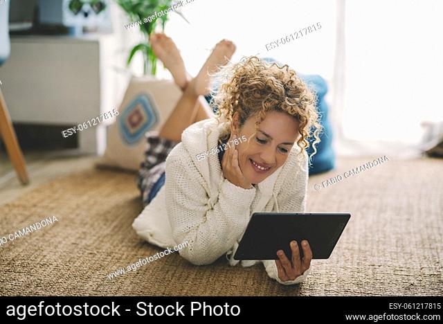 Happy adult woman laying on the carpet floor at home using modern tablet and internet connection wireless at home. Female people enjoy indoor relax leisure...