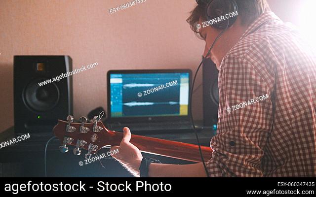 Young musician composes and records music playing the electric guitar, using computer and keyboard, from the front view