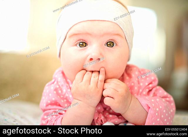 Cute 6 months old Baby girl infant on a bed on her belly with head up and fingers in mouth, looking towards camera with her big eyes