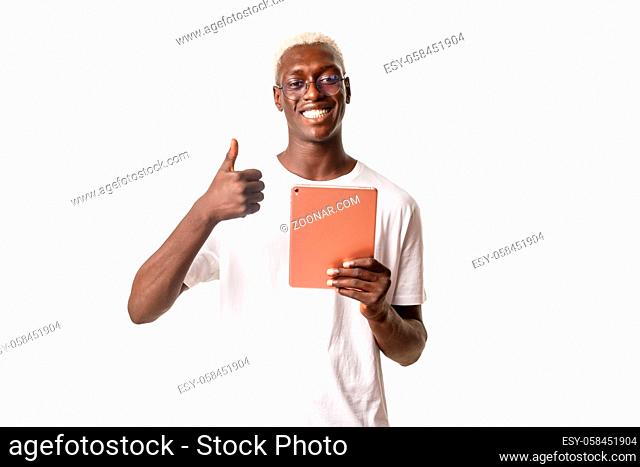 Portrait of happy good-looking african american blond male student, guy in glasses showing thumbs-up while using digital tablet, white background