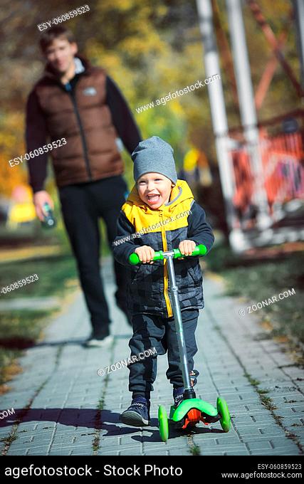 A happy little boy on a scooter walks in the park with his father on an autumn day. The child rides a scooter on the sidewalk and the parent walks behind and...
