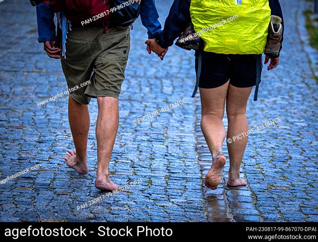 17 August 2023, Mecklenburg-Western Pomerania, Rostock: Barefoot vacationers walk in the rain on a cobbled street in the Baltic Sea resort of Warnemünde